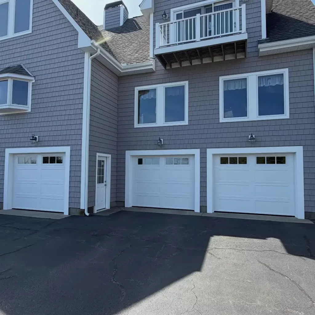 Three new white residential garage doors installed on a gray shingle home in Westerly, Rhode Island, showcasing professional garage door installation services.